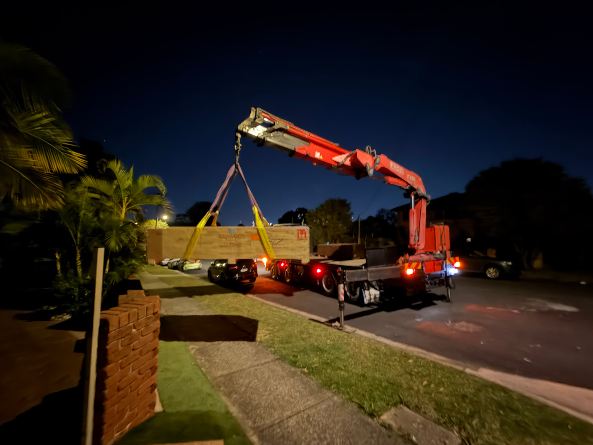 Everest Transport crane truck with outriggers set, arm extended, lifting a long timber crate at dusk