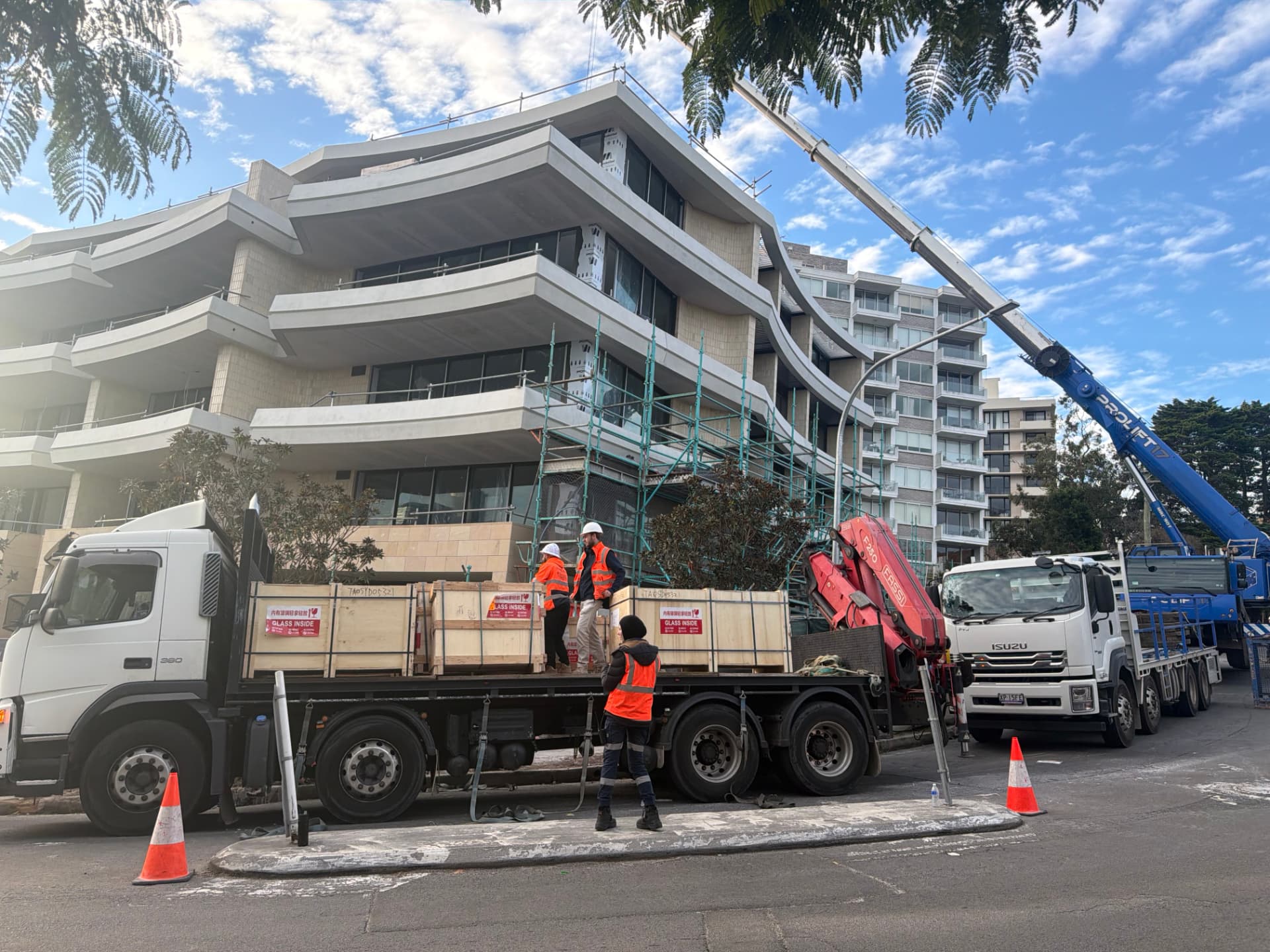Two crane trucks delivering crated glass panels beside a scaffolded apartment building under construction