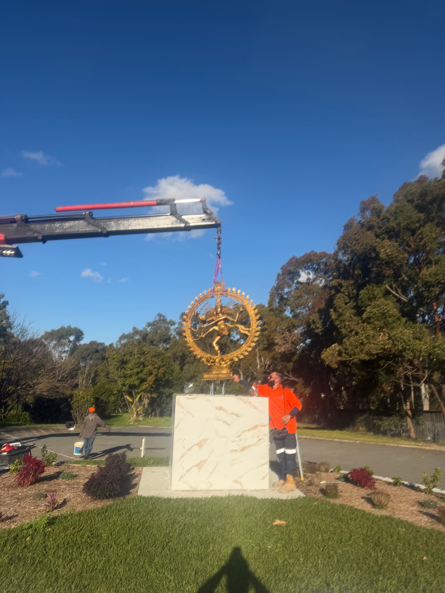 Crane arm lowering a gold statue onto a tiled plinth in a landscaped garden