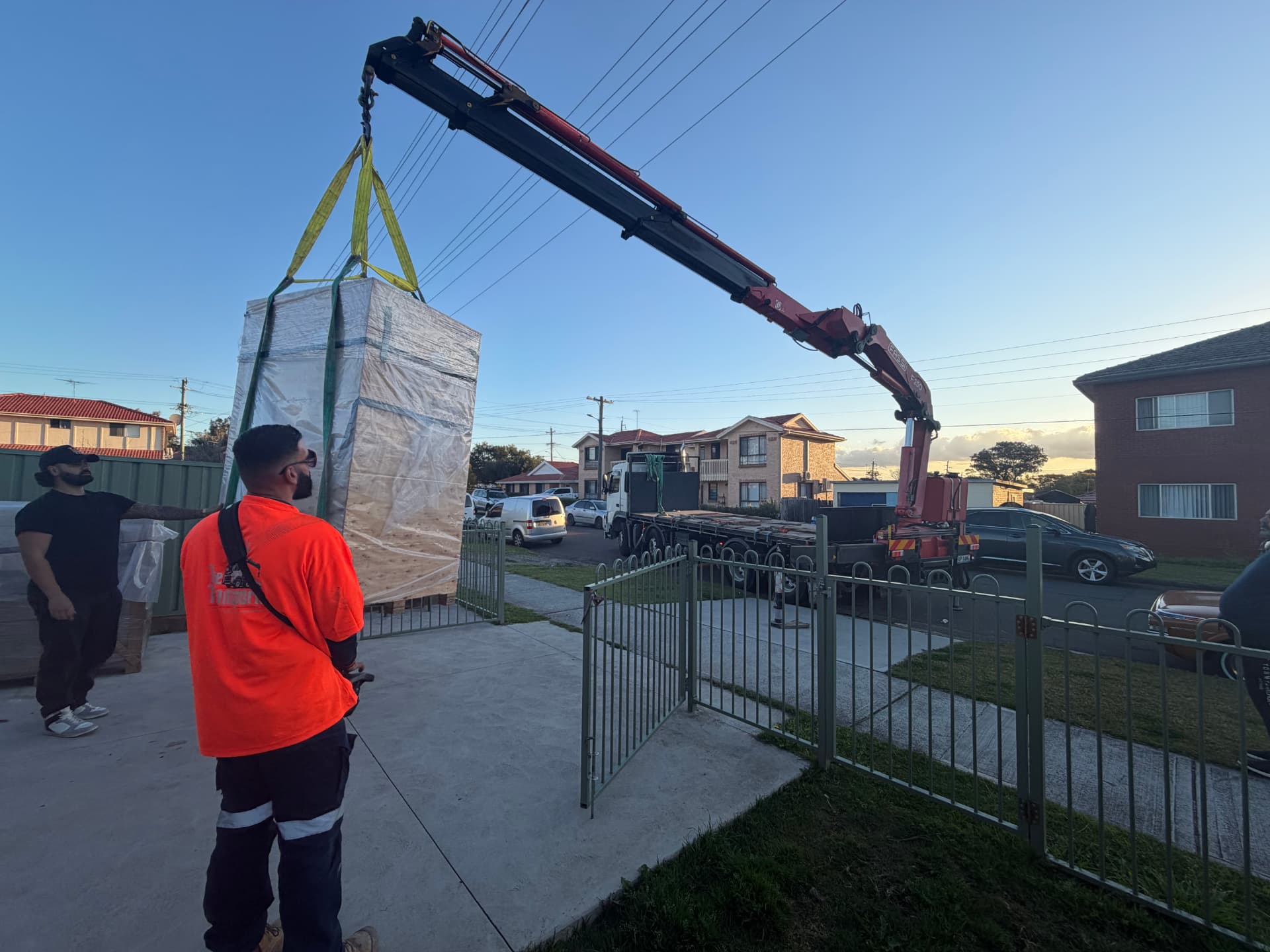 Crane truck lifting a large wrapped item over a residential steel fence in suburban Sydney