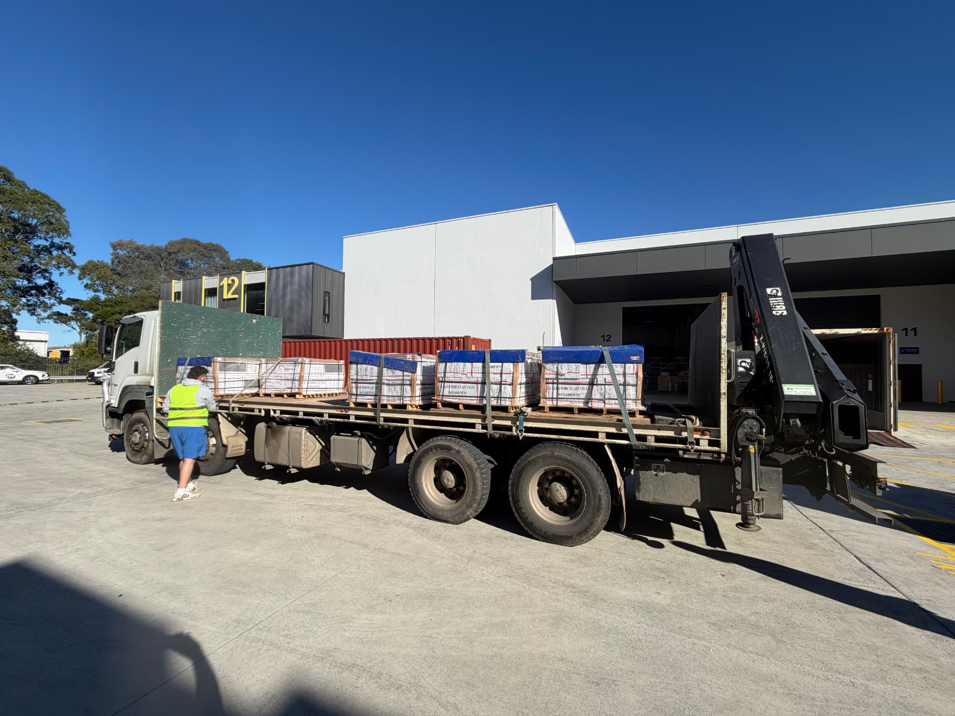Flatbed crane truck loaded with strapped pallets of glazed porcelain tiles at a warehouse loading dock